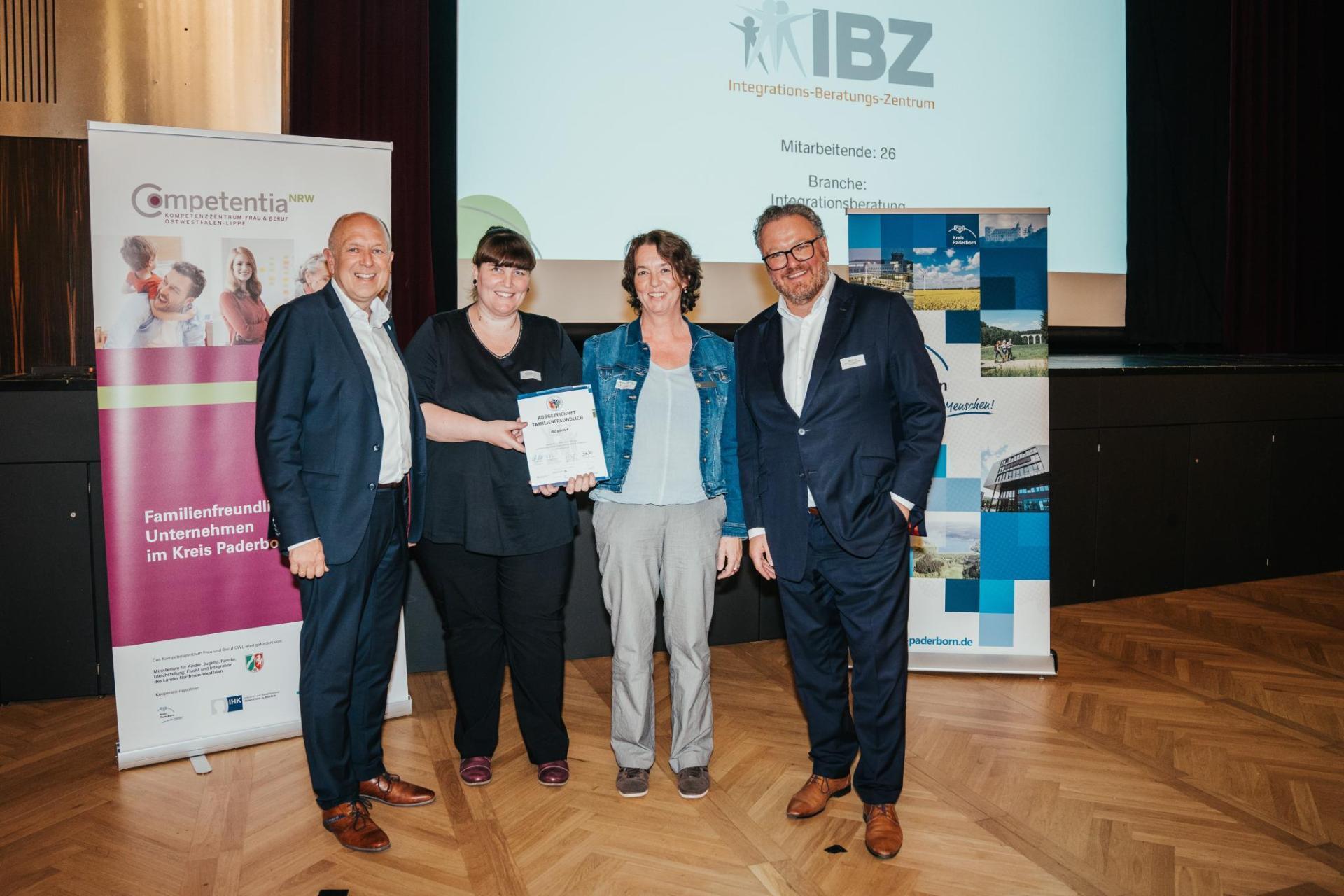 Vier formal gekleidete Personen stehen auf einer Bühne, auf der ein Banner für "IBZ" und eine Präsentationsleinwand mit den Angaben "Mitarbeiten: 26" und "Branche: Integrationsberatung".